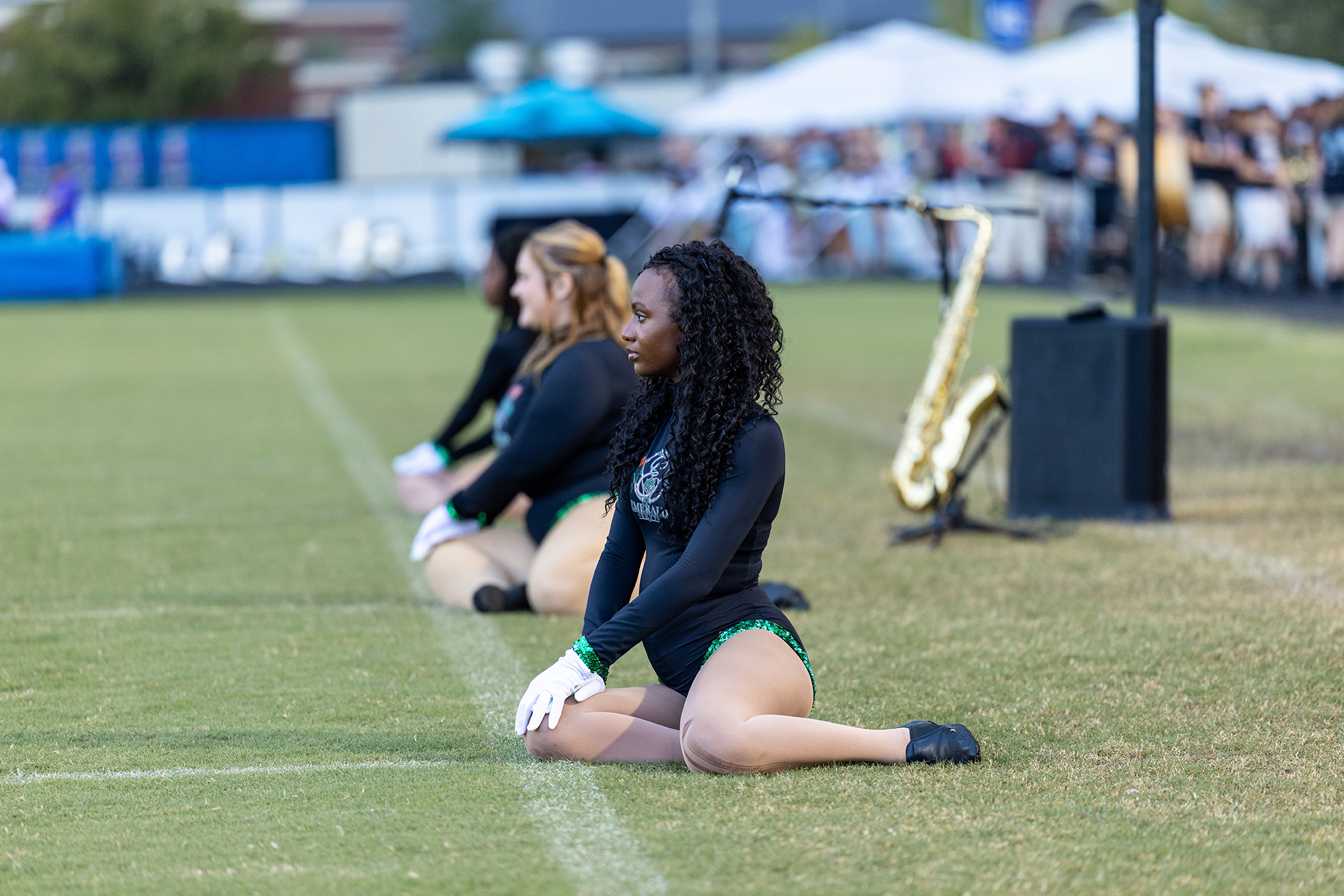 Two cheerleaders in uniform sitting on a field, one in front wearing white gloves. A saxophone stands nearby.