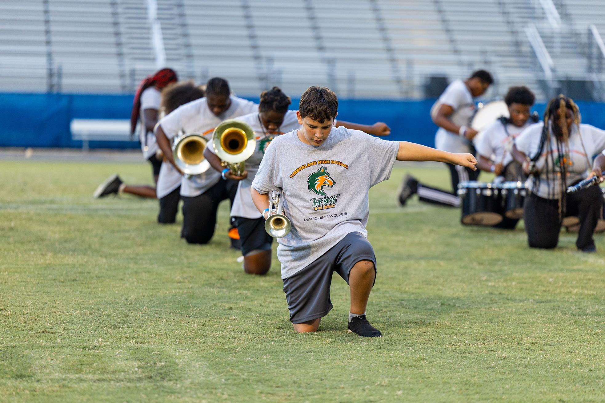 A group of musicians in white shirts and gray shorts performs on a field, with instruments in hand.