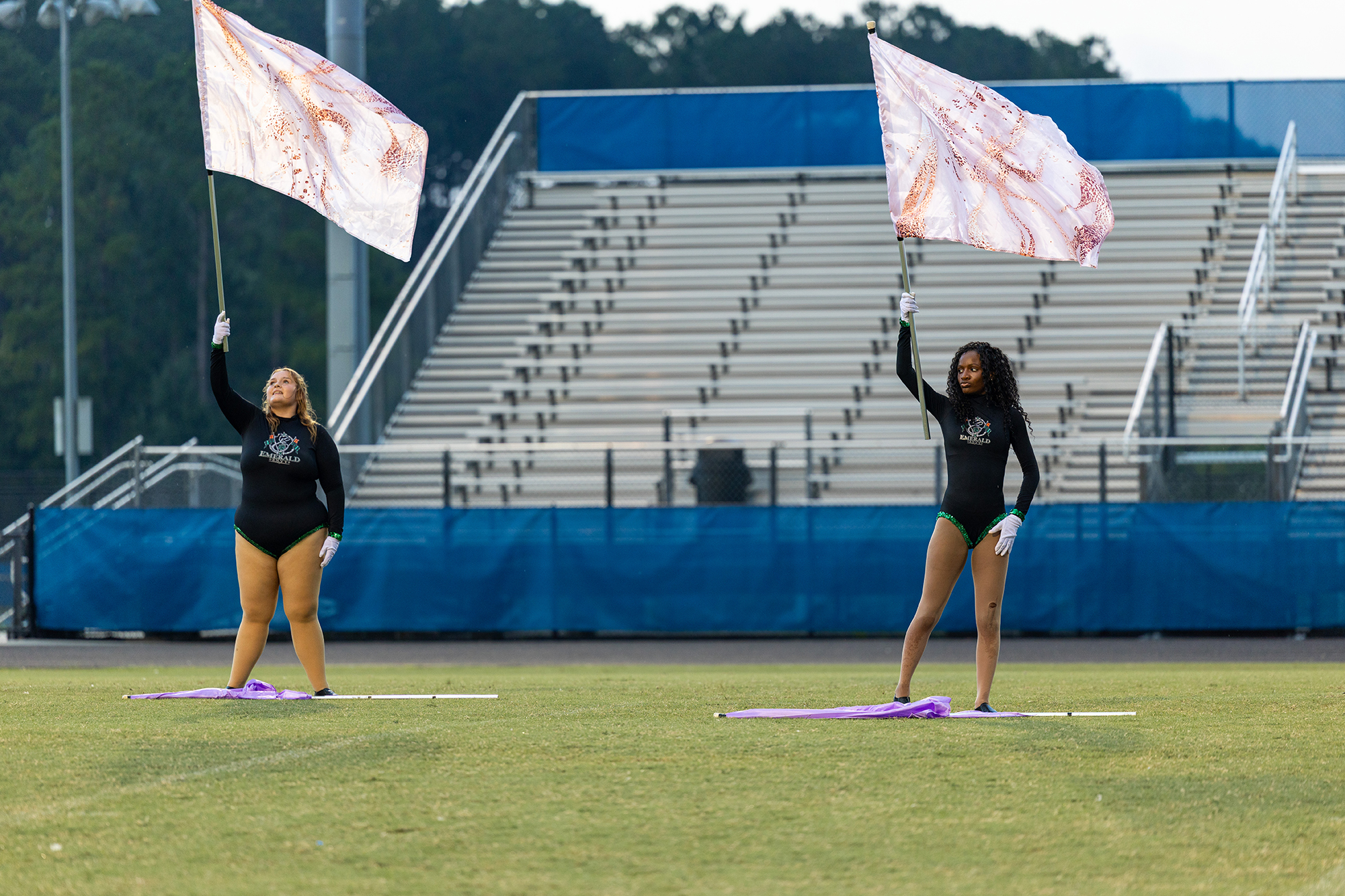 Two women holding flags in front of a partially empty stadium. One woman is wearing a black uniform, the other a green one.