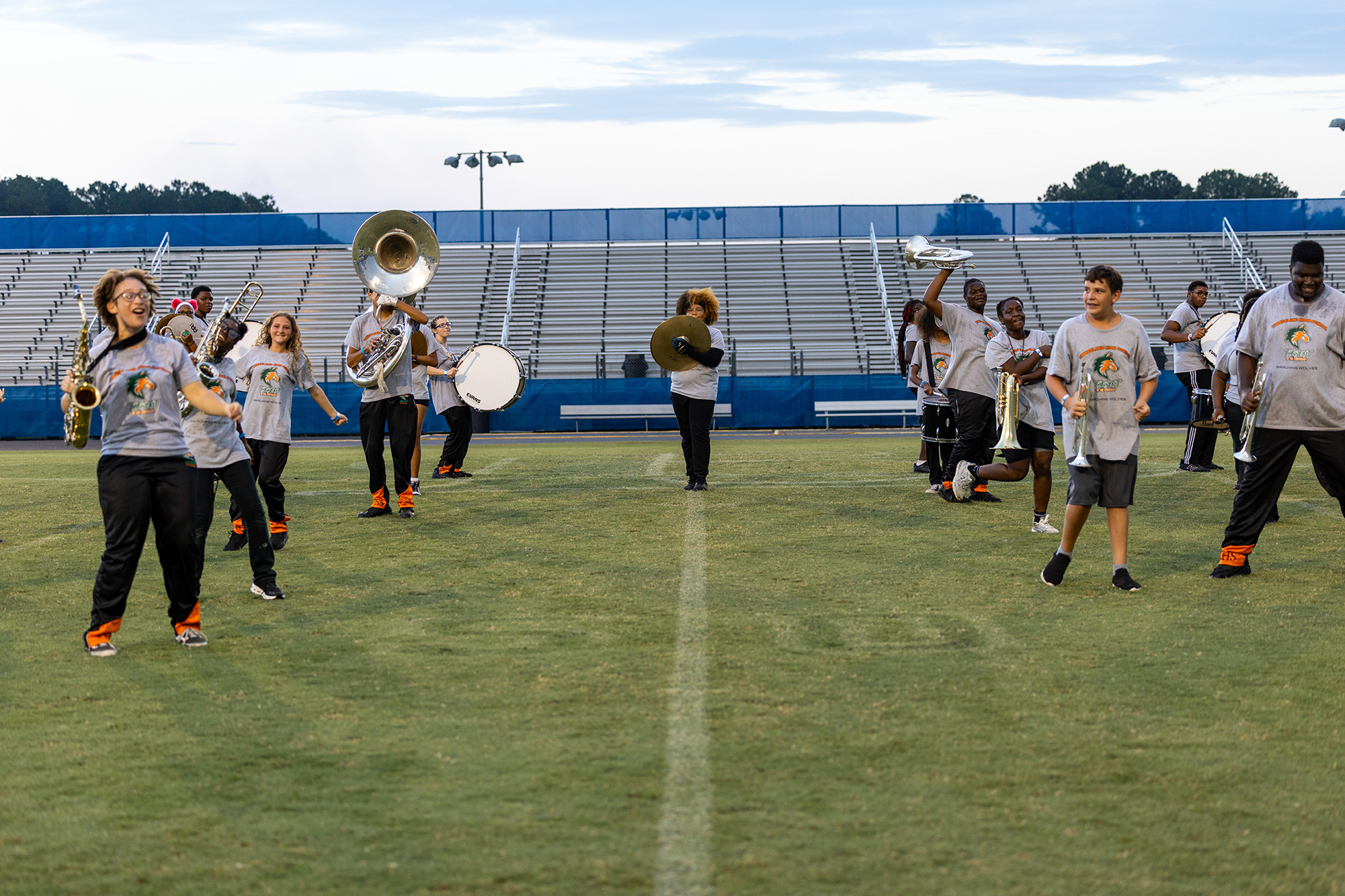 A band practices on a field. Members hold instruments, some walk while others stand. A stadium with bleachers is in the background.
