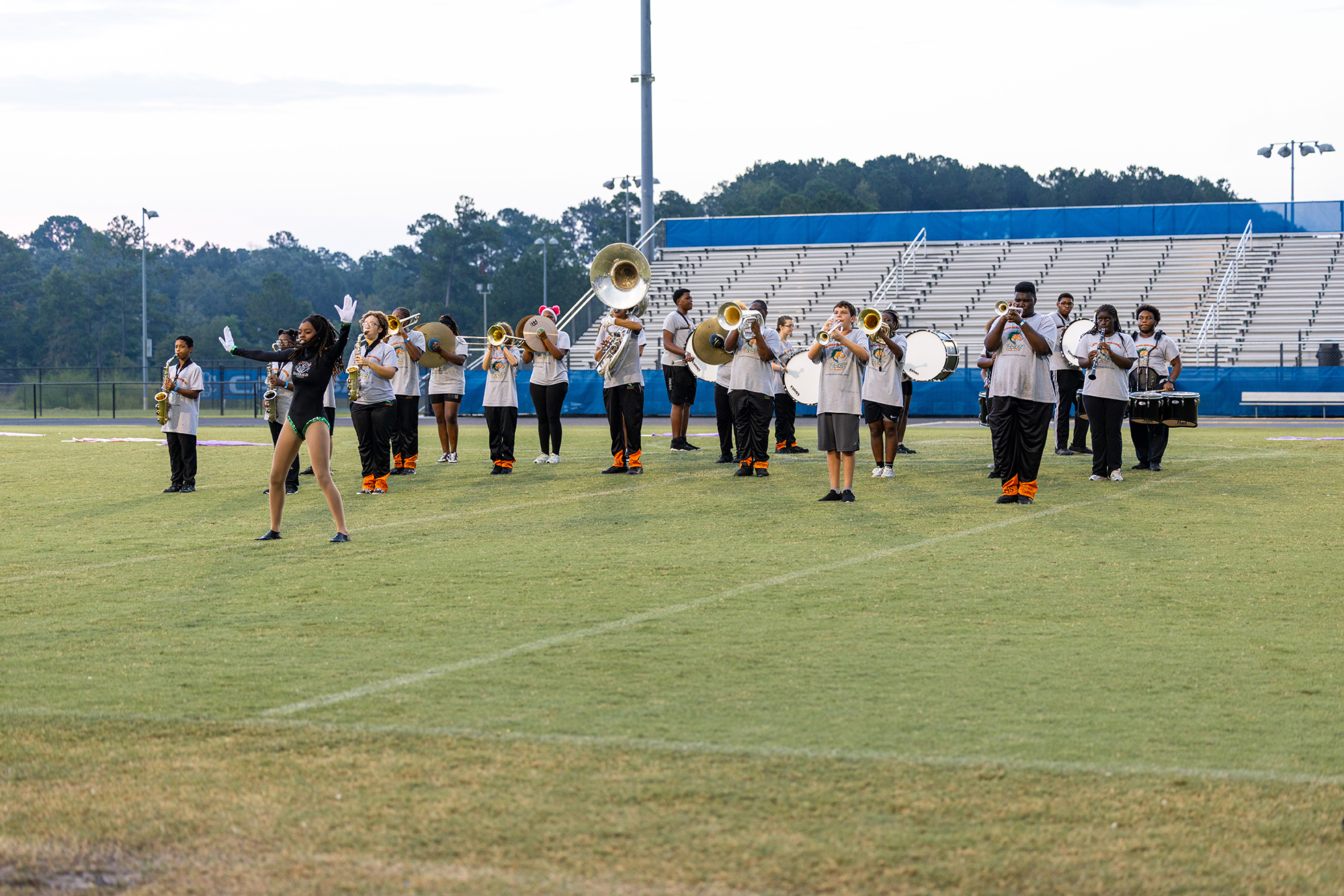 A band practices on a football field with a bleacher background. Some members play instruments while one person performs a cartwheel.