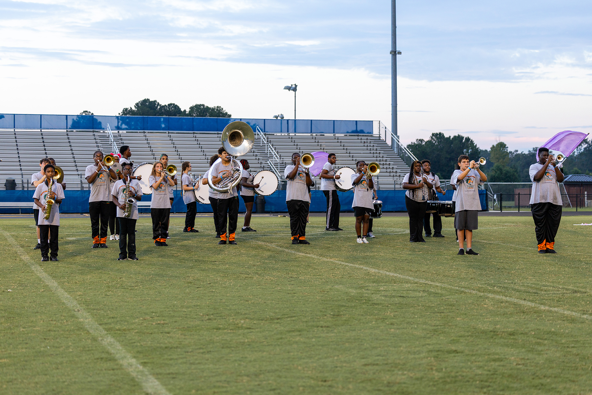 A marching band plays instruments on a football field, with bleachers and trees in the background.
