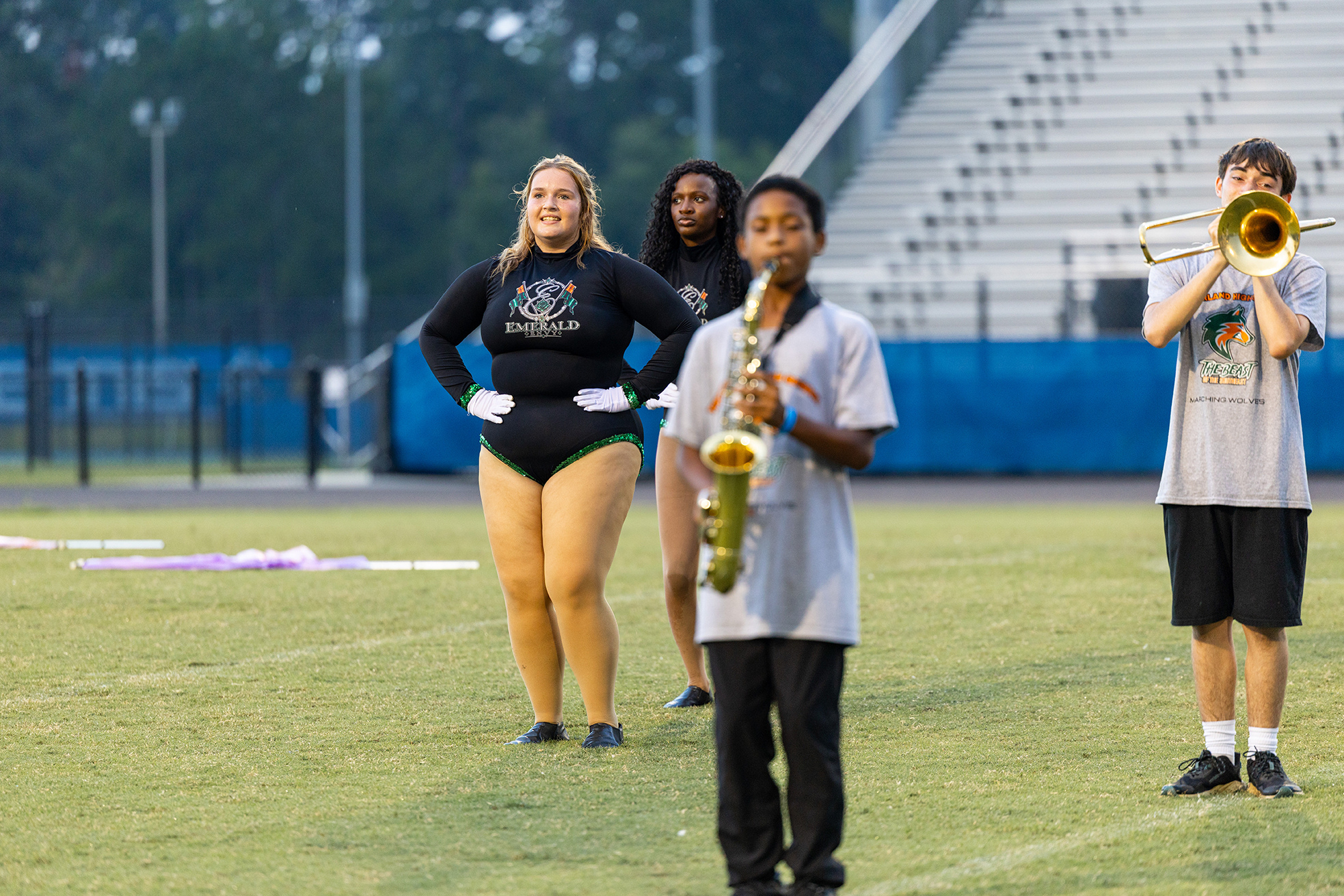 Four individuals stand on a grass field. A woman and a boy play musical instruments. An empty stadium is in the background.