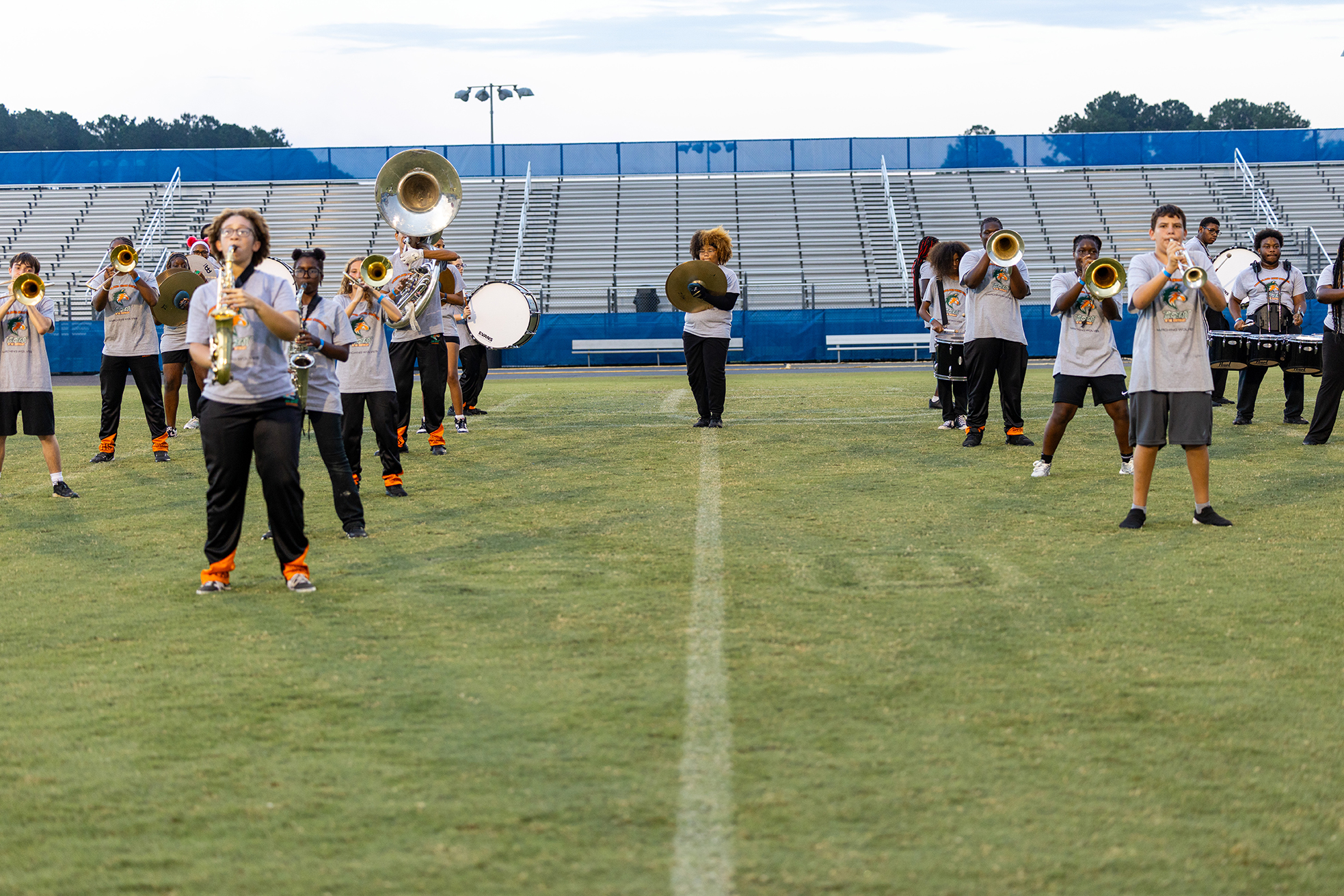 A group of musicians plays instruments on a field. They stand in a row, some holding trumpets, while empty bleachers are in the background.