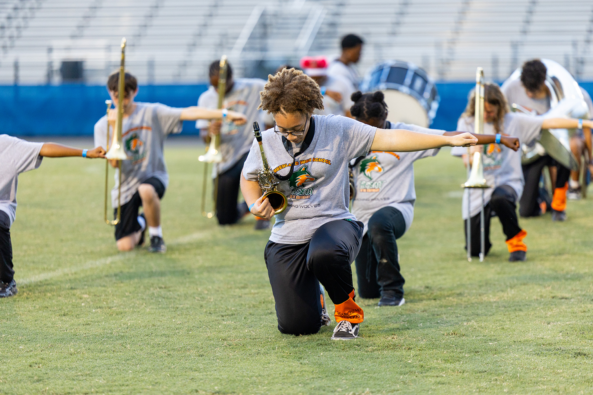 A group of band members on a green field, some kneeling, holding trumpets, with empty bleachers in the background.