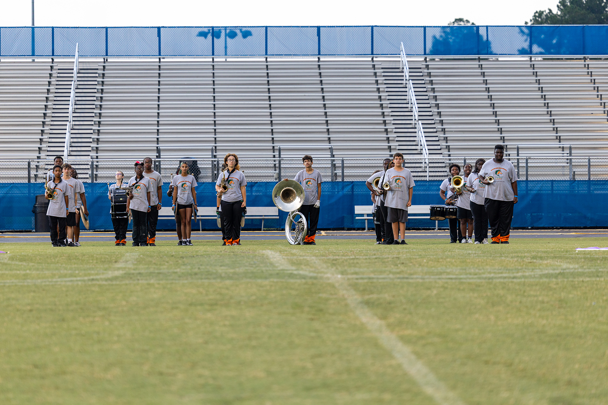 A band stands on a field, holding instruments. A few are seated. Behind them, an empty bleacher.