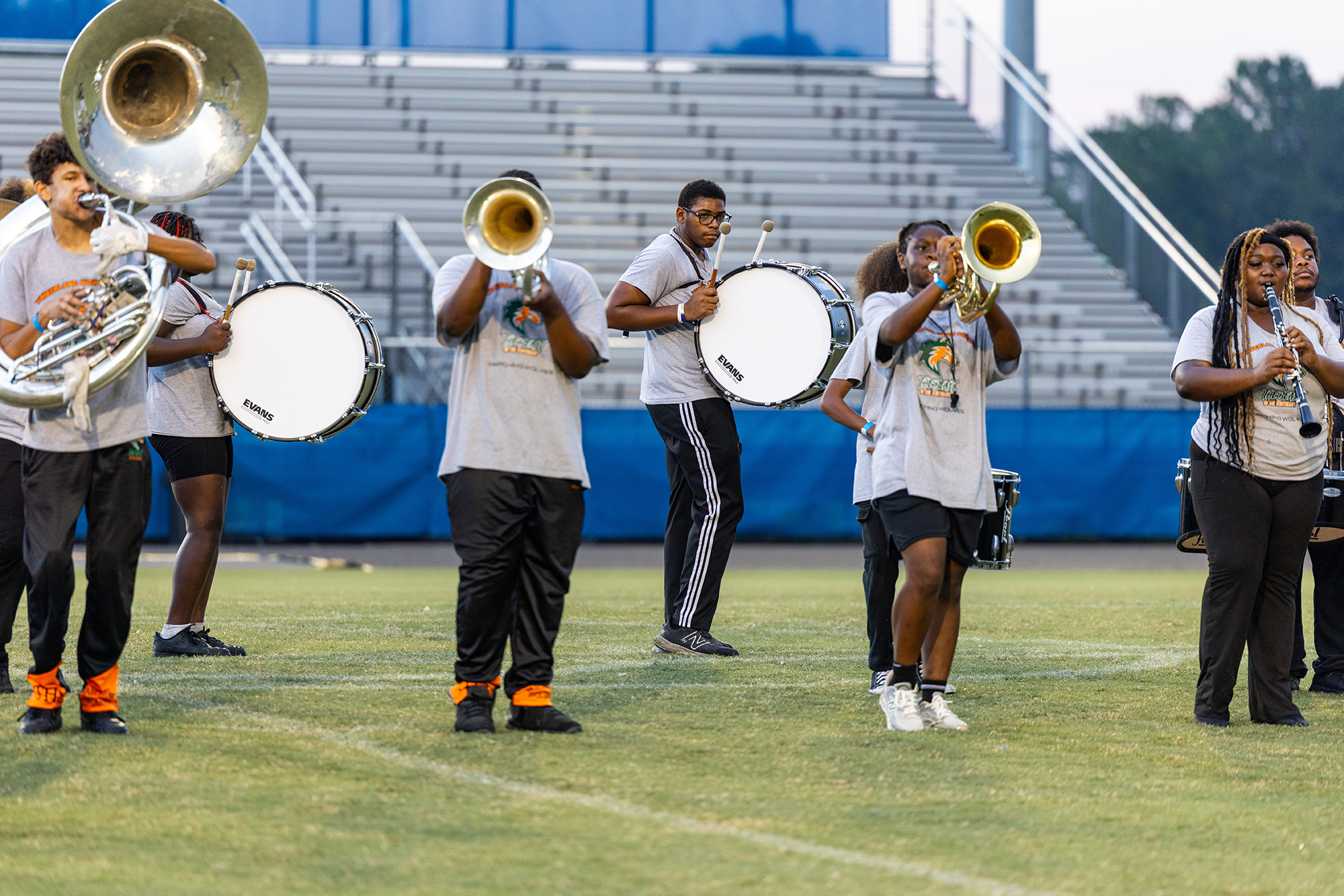 A band performs on a field, with players holding brass instruments. Empty bleachers are in the background.