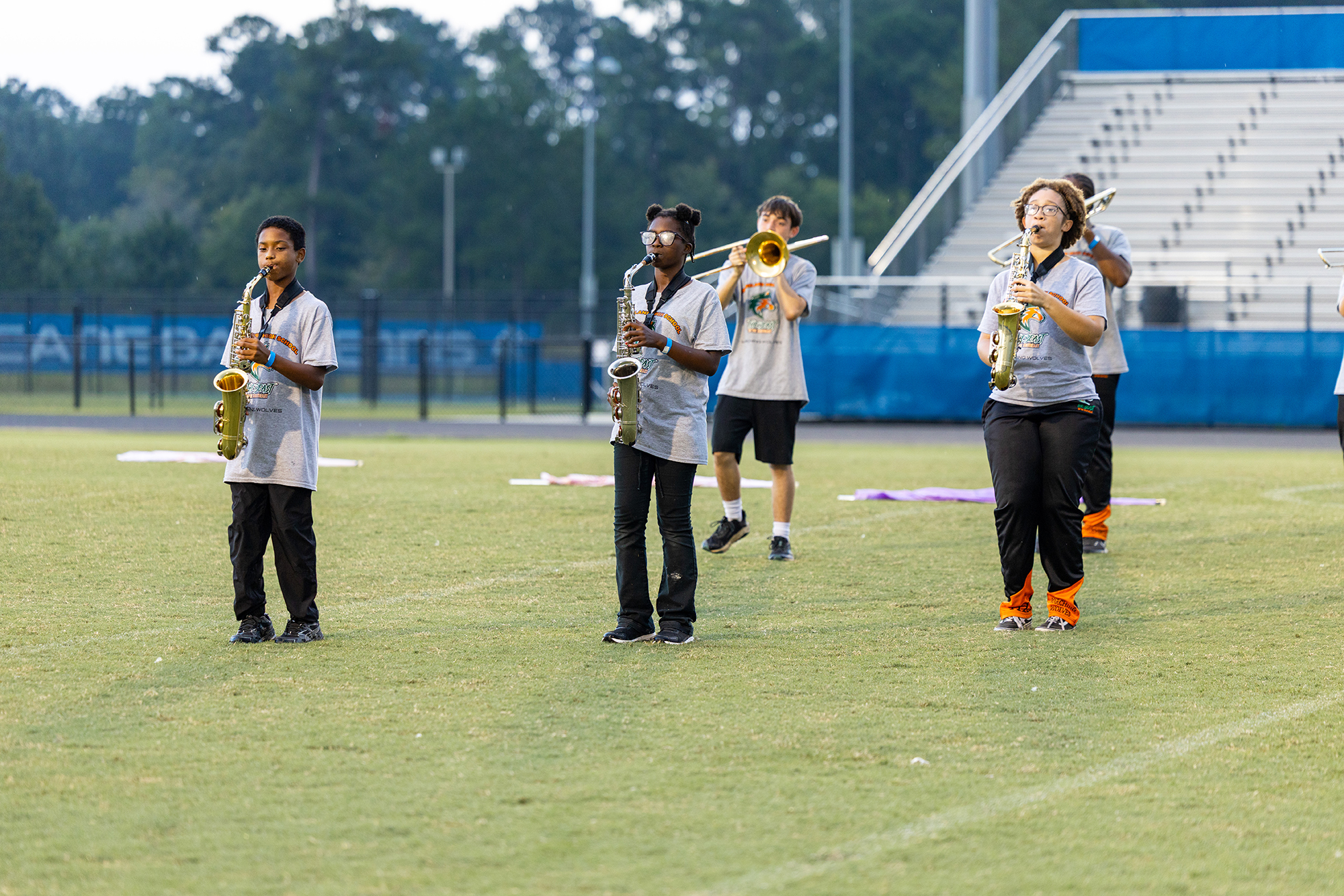 Five individuals on a field holding musical instruments. Two in front, one with a saxophone, one with a trumpet.