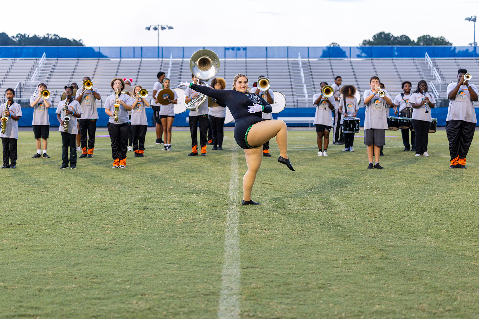 A person kicking a leg high while other band members play instruments on a field with stadium seating.