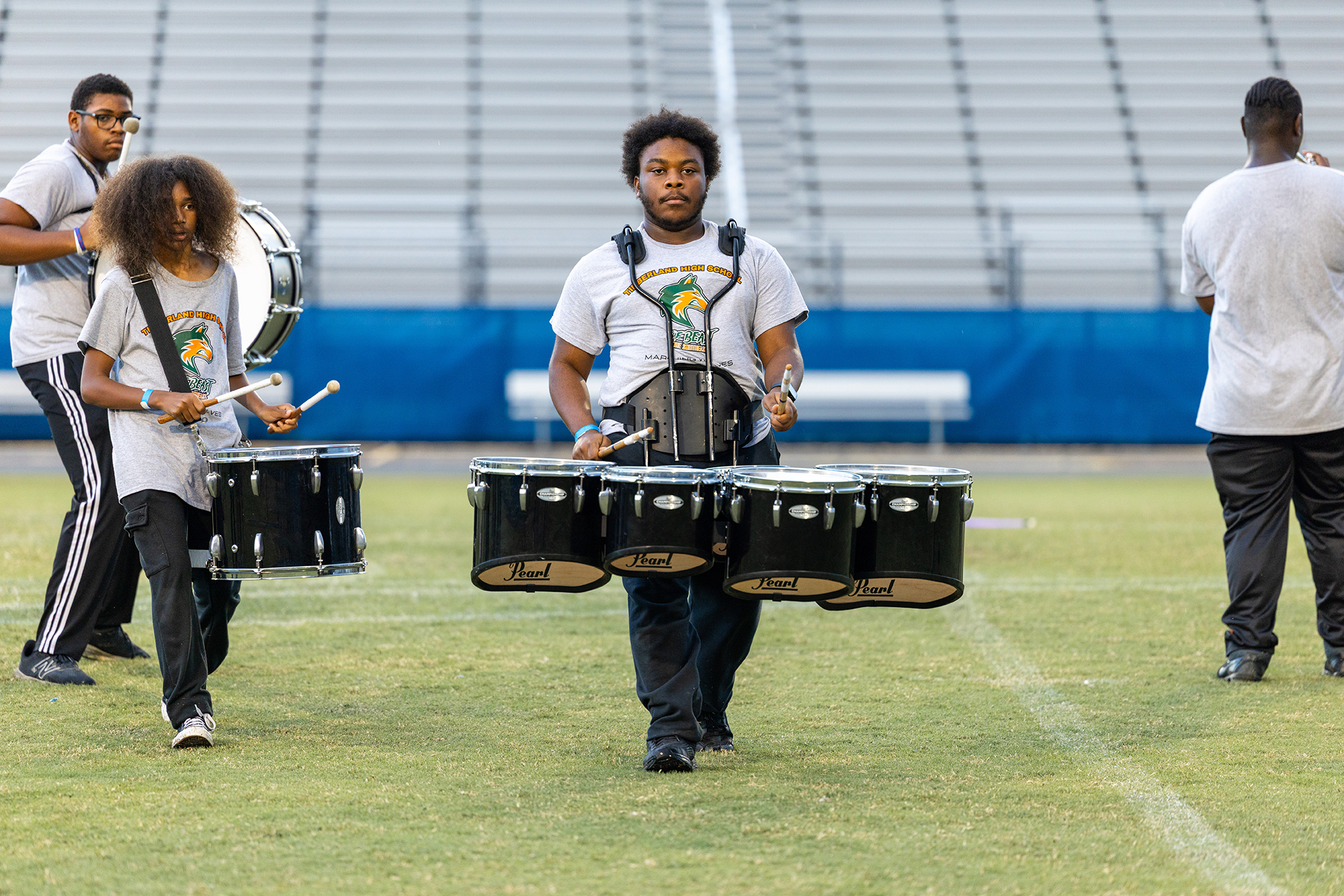 Four individuals wearing white shirts and carrying drums, performing on a football field. Empty bleachers in the background.