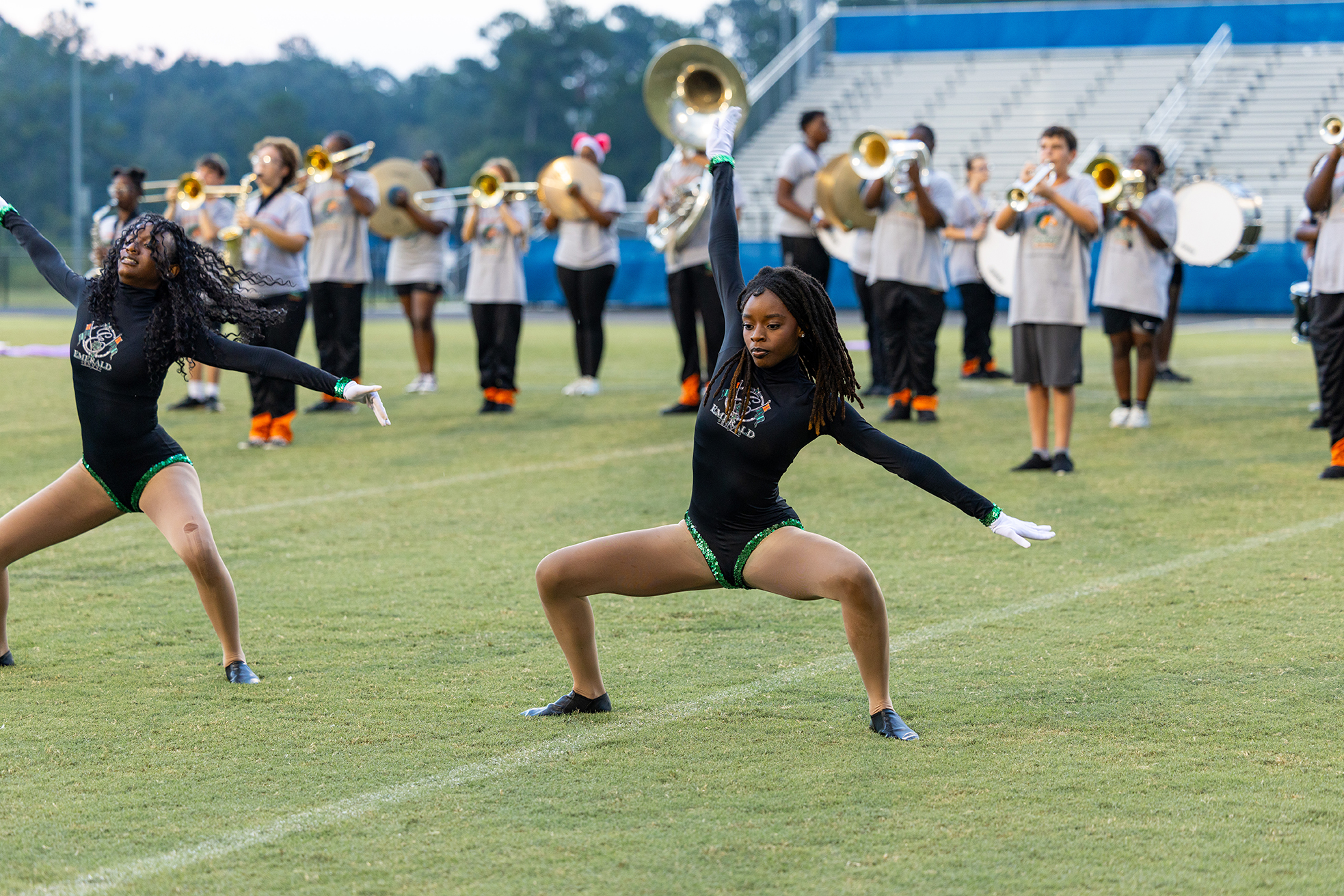 Two cheerleaders performing a split on a field with a marching band in the background.