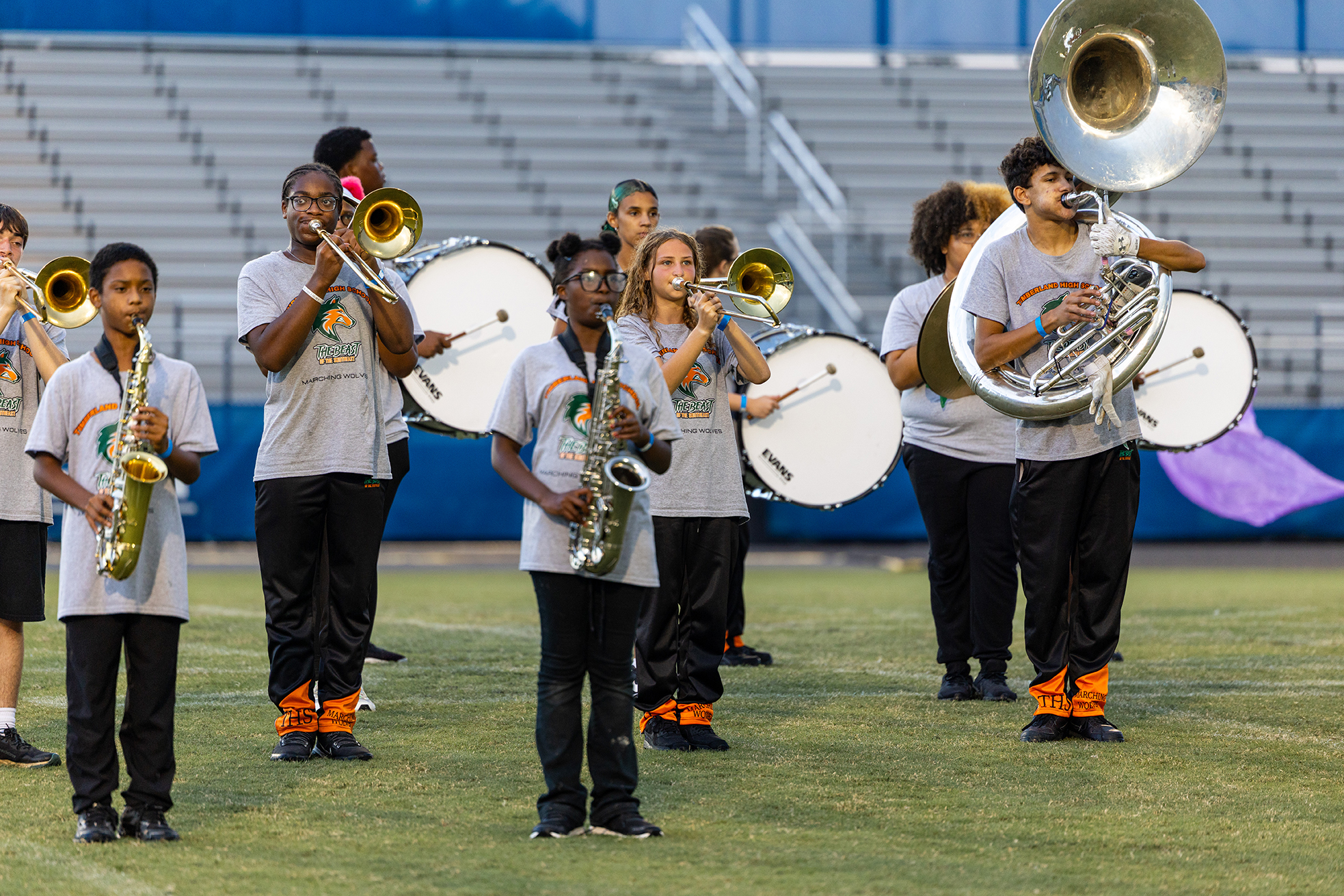 A school band performs on a field with instruments. They wear matching uniforms and stand in front of bleachers.