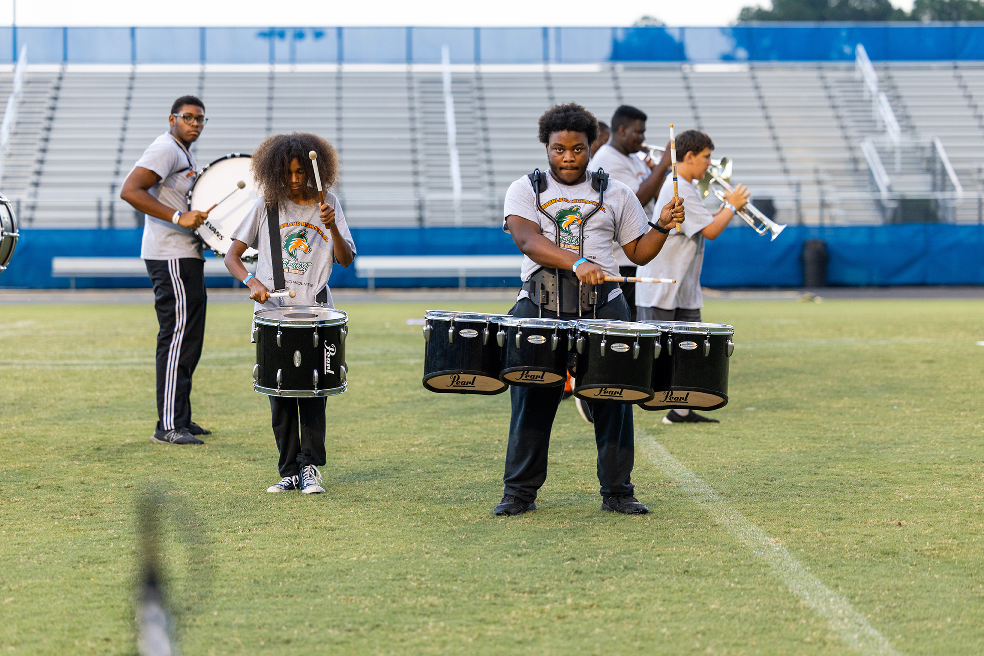 Group of musicians on a field. Two play drums, two play trumpets. Empty bleachers in the background.