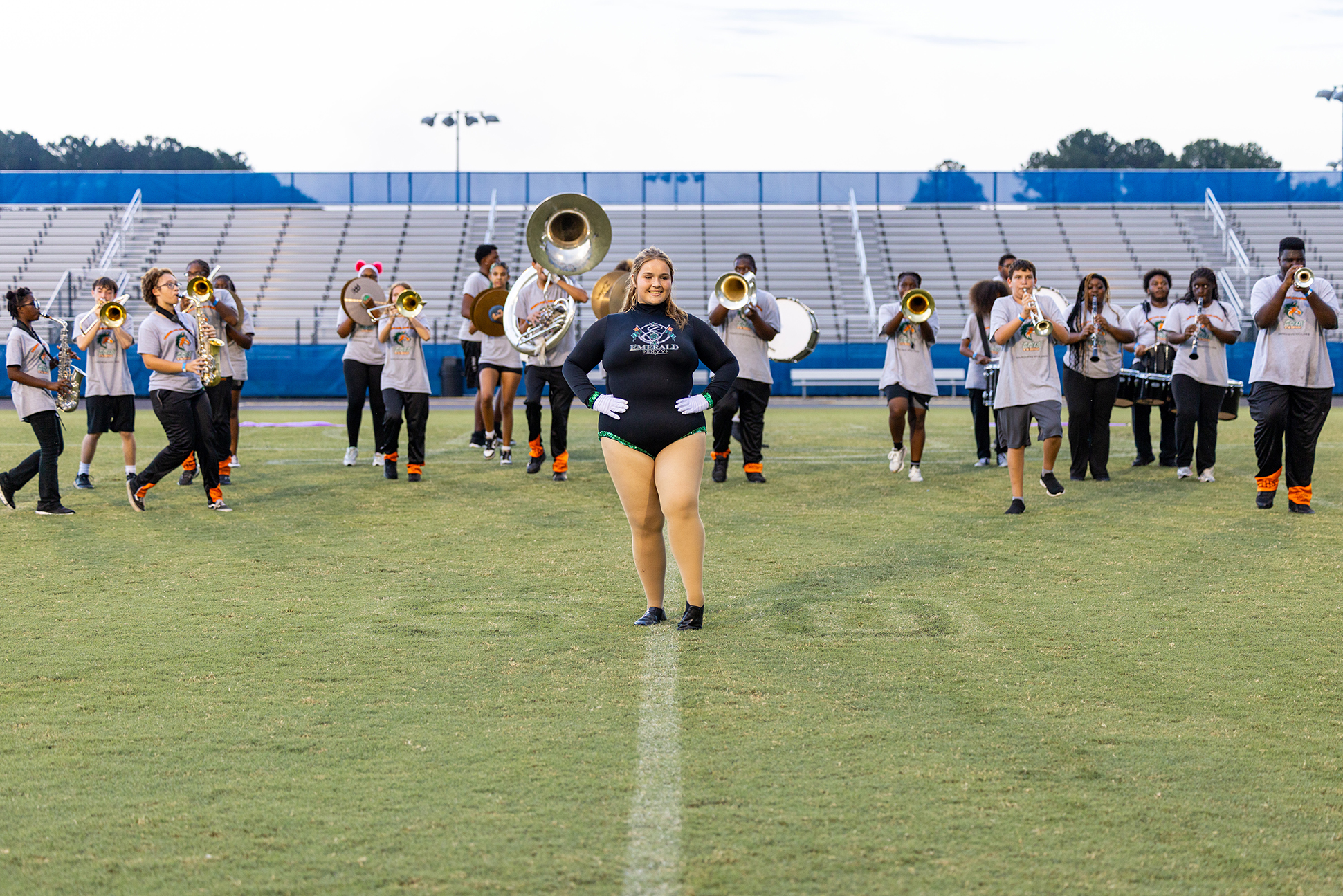 A group of musicians with brass instruments stands in a line on a grassy field. Some wear white uniforms.