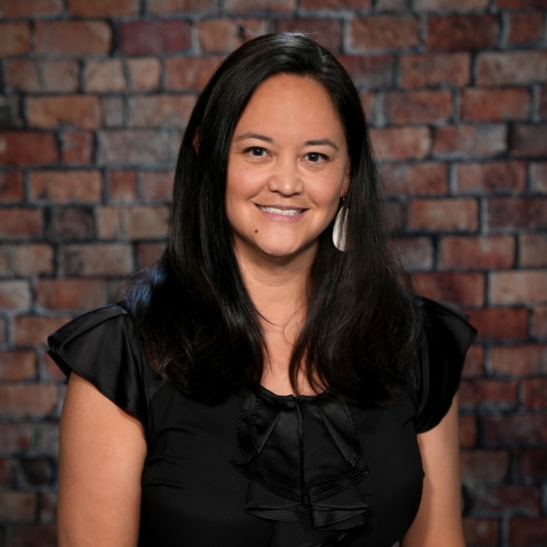 A smiling woman with long dark hair stands in front of a brick wall, wearing a black ruffled blouse.