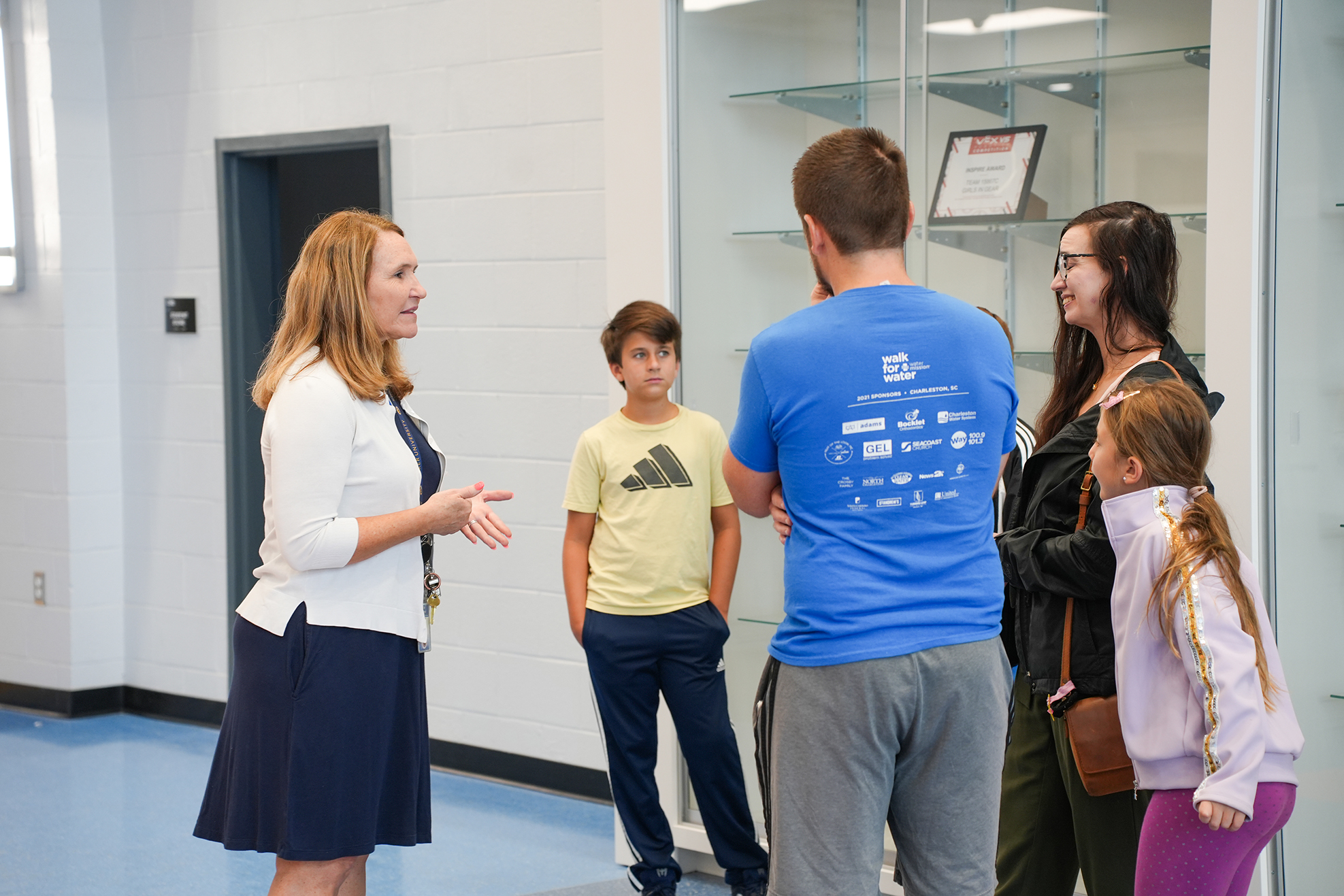 Five people in a room with a white wall and blue floor, standing and talking.