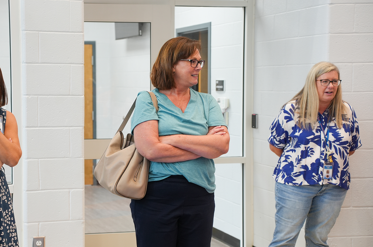 Three women stand in a hallway, two facing each other, one with arms crossed. The middle woman holds a bag.