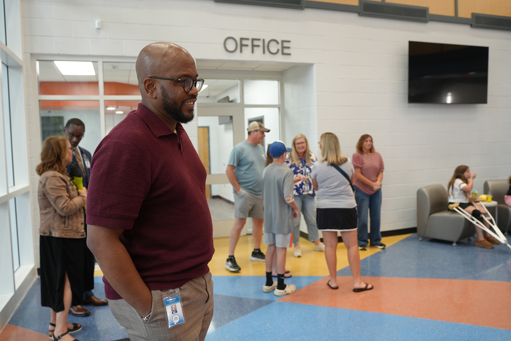 A man stands in a lobby with other people. He wears glasses and a maroon polo shirt. Behind him is a sign that reads "OFFICE".