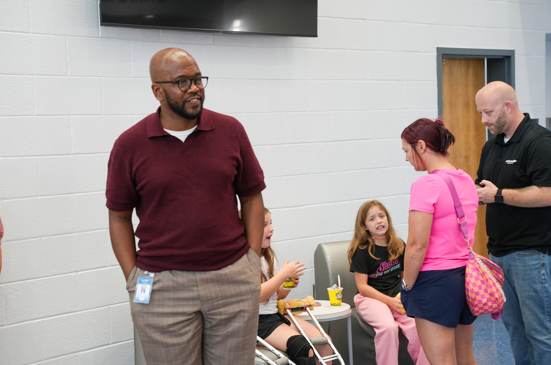 Several adults and children in a room. A man in a maroon shirt stands with his hands on his hips.