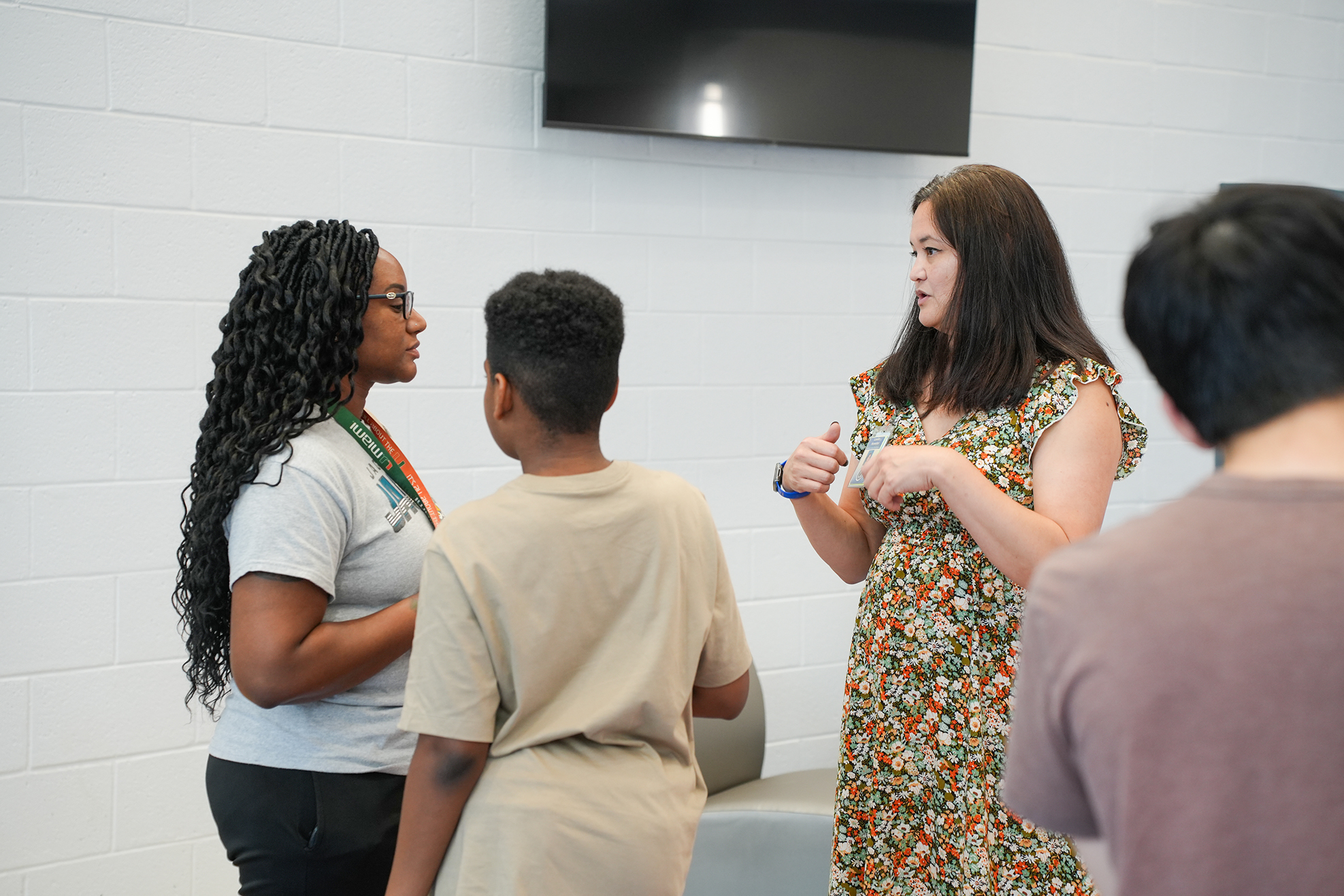 Four people converse near a white wall. A woman in a floral dress addresses a boy, while another woman listens.