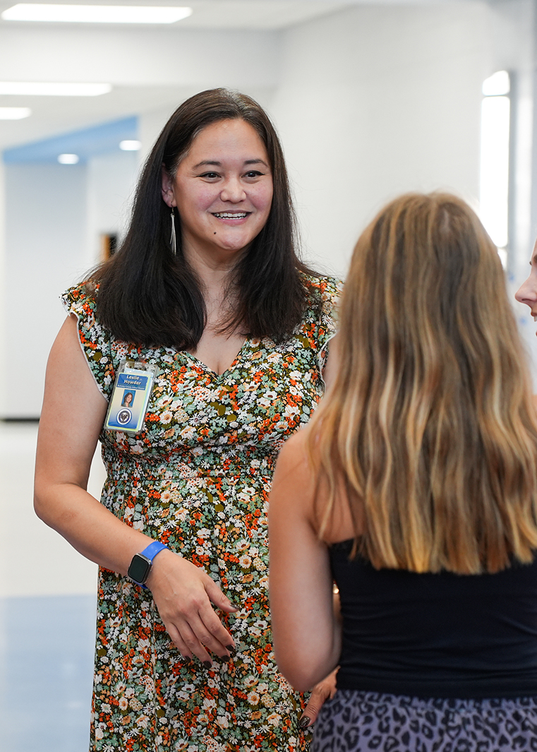 Woman in floral dress stands smiling, talking to a person in a black tank top. Background shows a well-lit hallway.