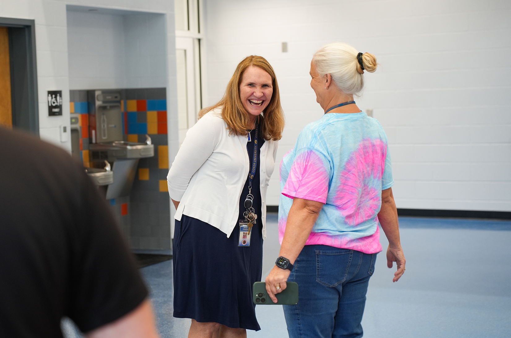 Two women converse in a hallway, one wearing a white cardigan, the other a tie-dye shirt. A sink and tiles are in the background.