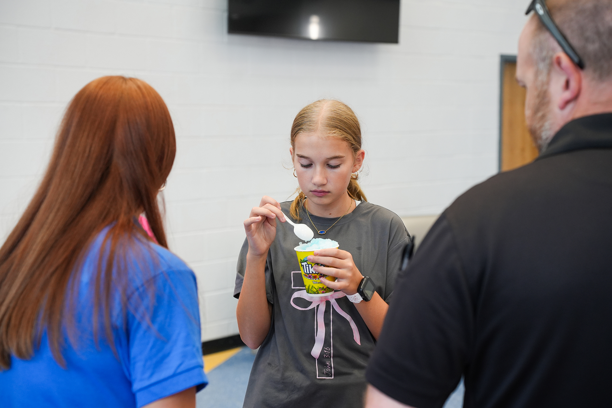 A young girl with blonde hair eats ice cream from a cup, while two adults watch.