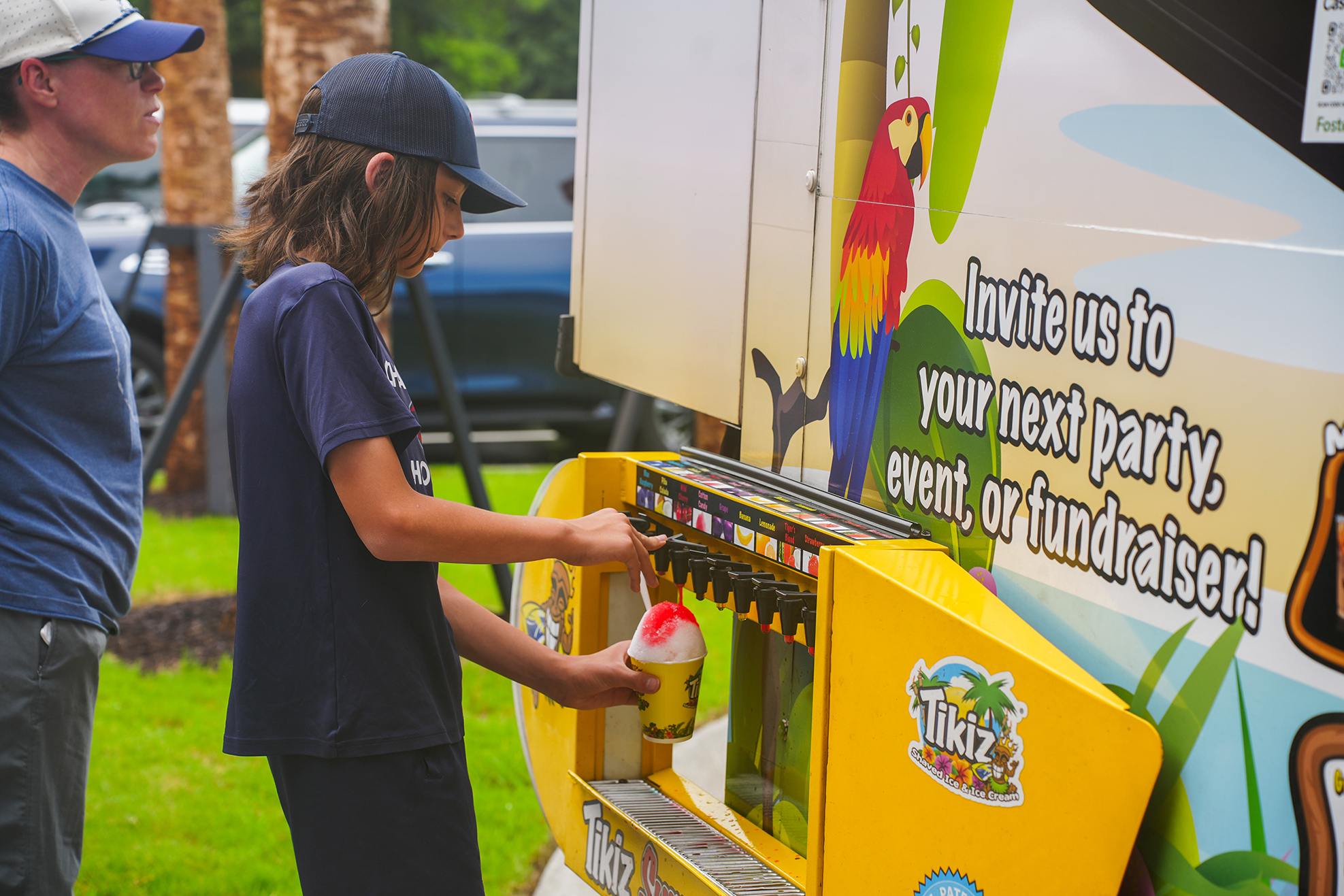 A person in a blue shirt and cap using a yellow drink dispenser outdoors. Another person in a blue shirt and hat stands nearby.