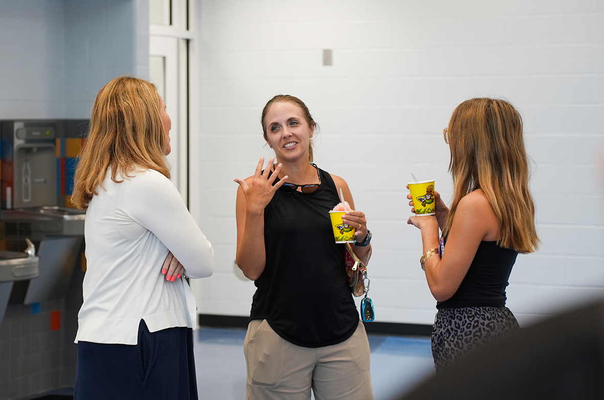 Three women conversing in a corridor with a wall and sink in the background.