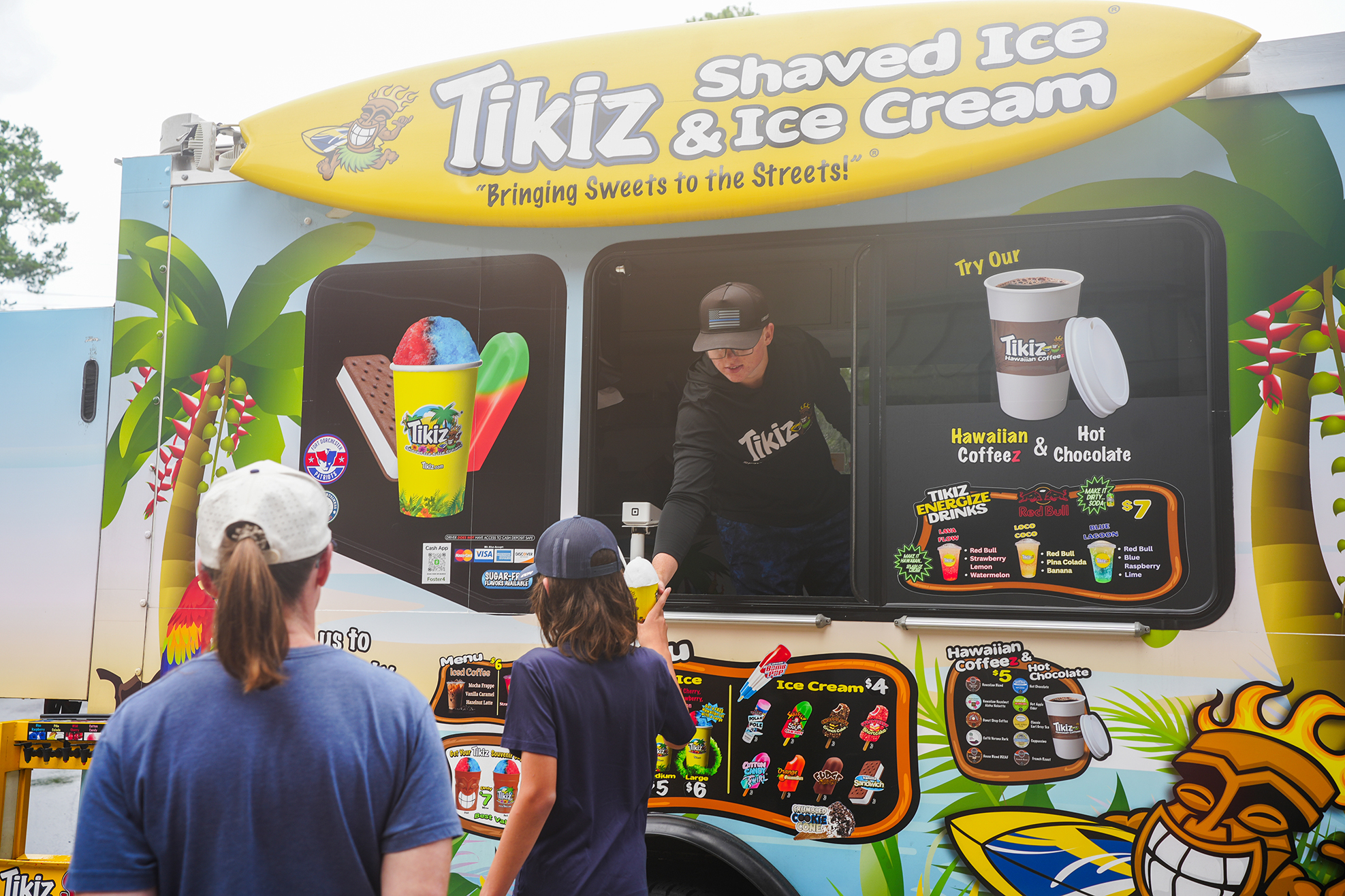 A shaved ice truck with the text "Tikiz & Ice Cream" on a yellow surfboard. Two people are looking at the menu.