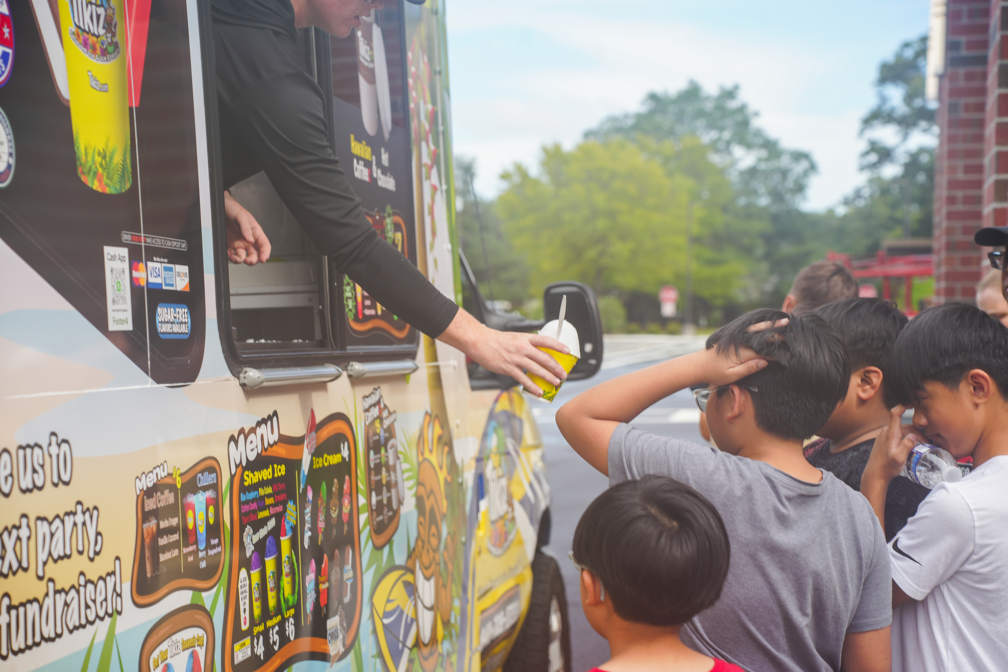 A person hands a cup out of a window of a food truck. Children gather to receive their orders.