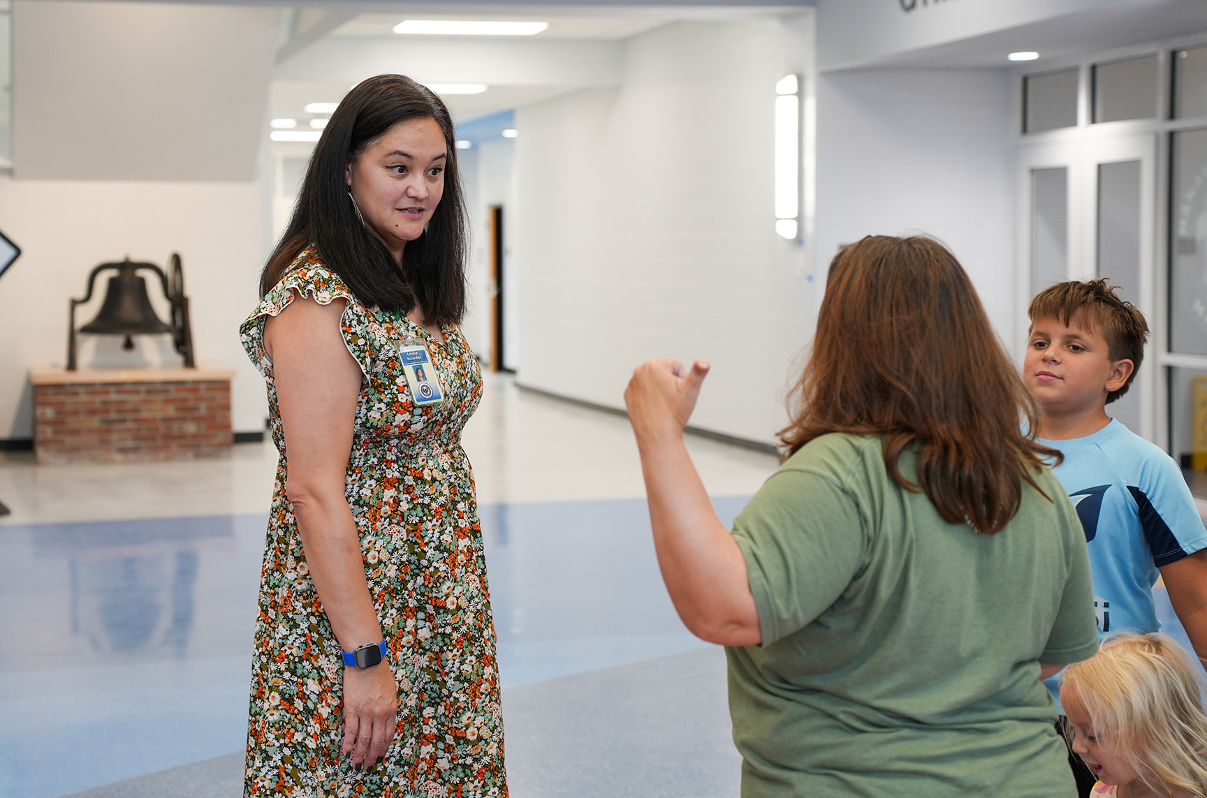 A woman speaks to another woman and two children. They are in a hallway with a bell and large windows.