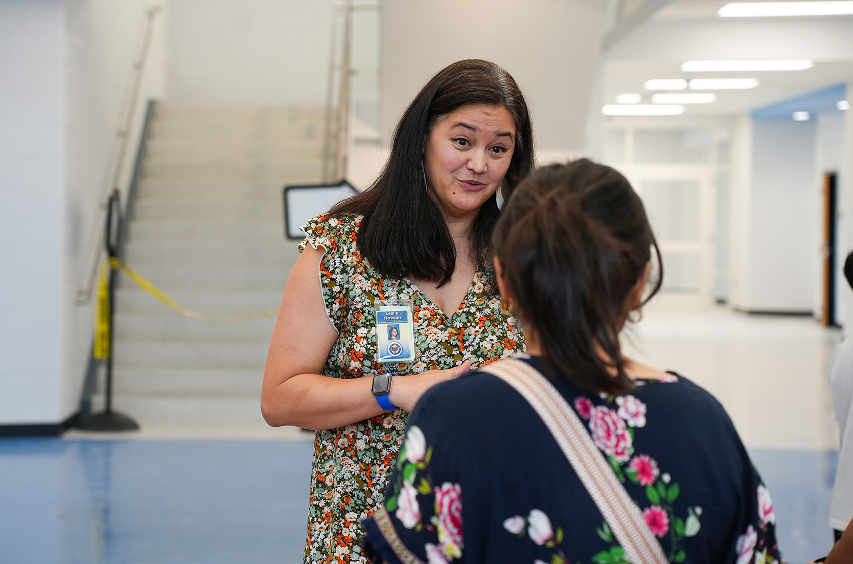 A woman with a badge speaks to another woman with a floral-patterned bag in a brightly lit hallway.
