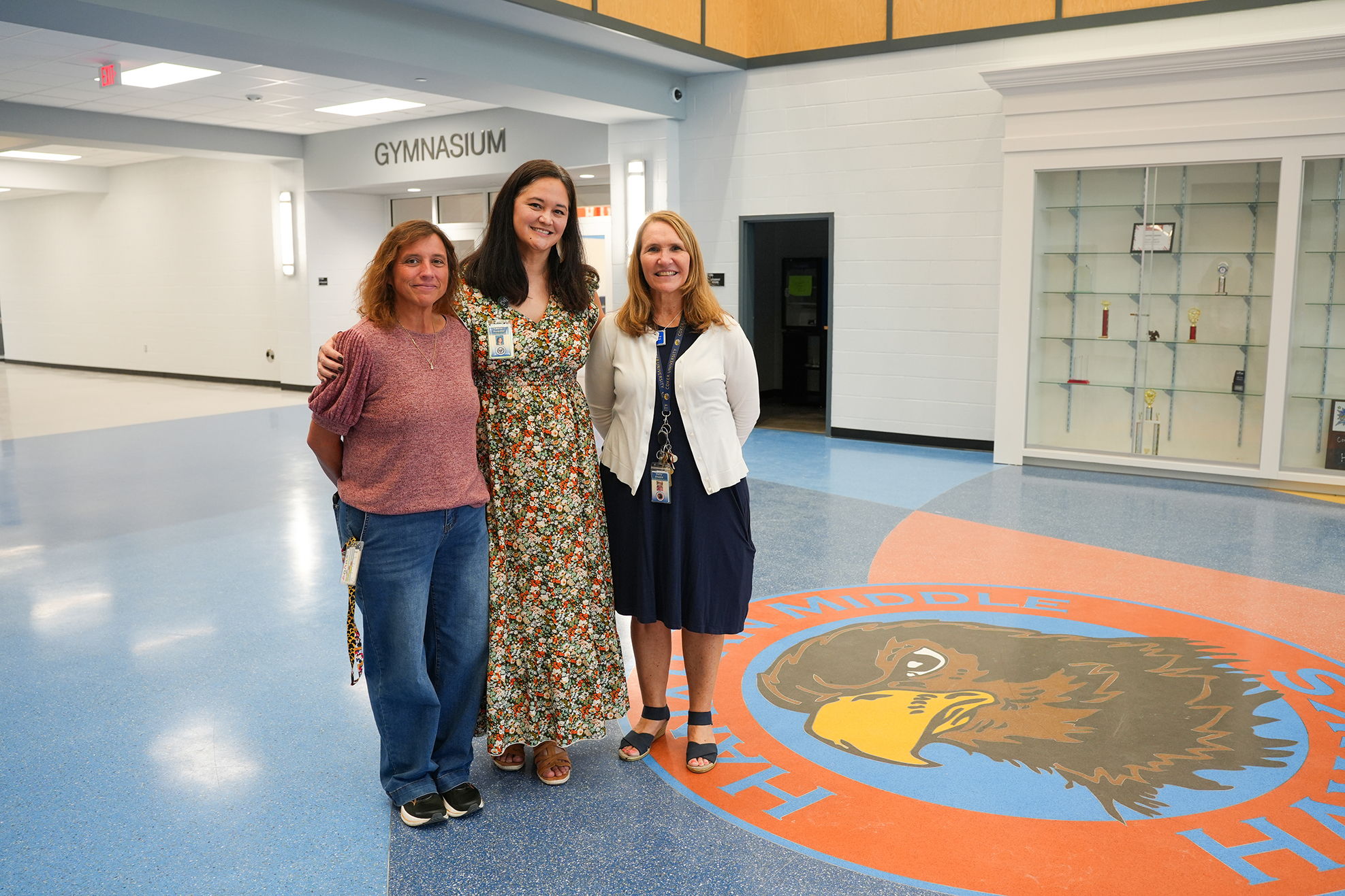 Three women in a hallway. Two wear dresses, one wears casual clothes. They stand near a floor design.