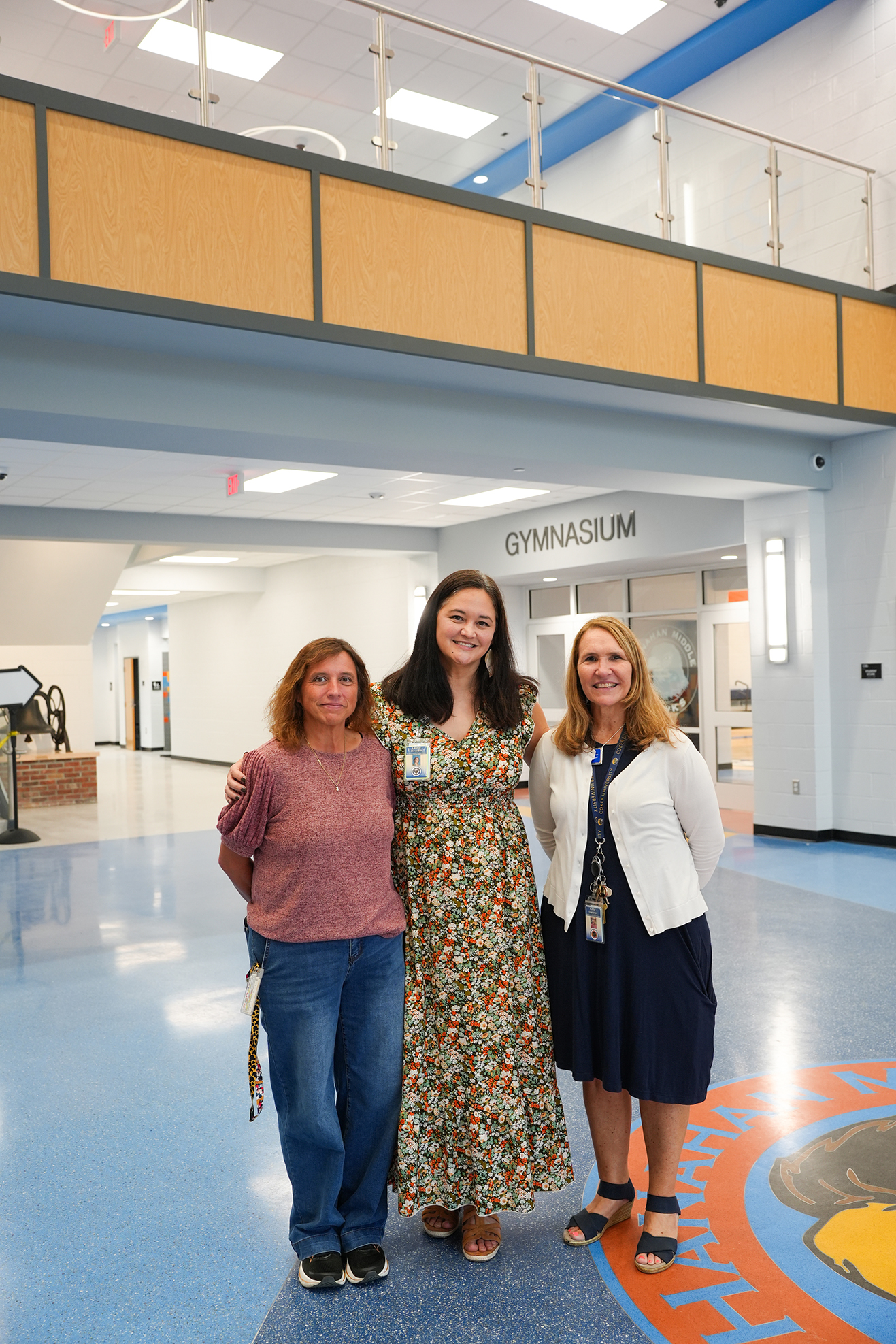 Three women pose for a photo in a school hallway with a blue floor. One wears a floral dress, another in a pink shirt, and the third a white jacket.