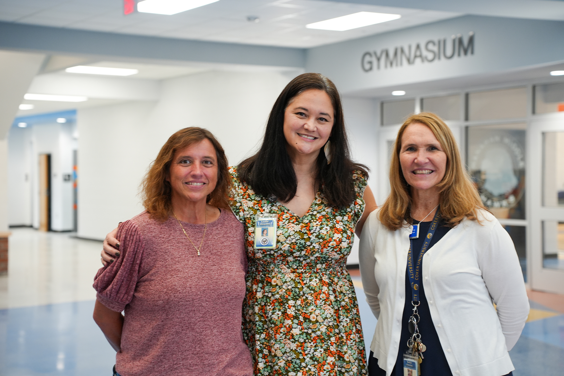 Three women standing together inside a building with a sign above them that reads "GYMNASIUM".