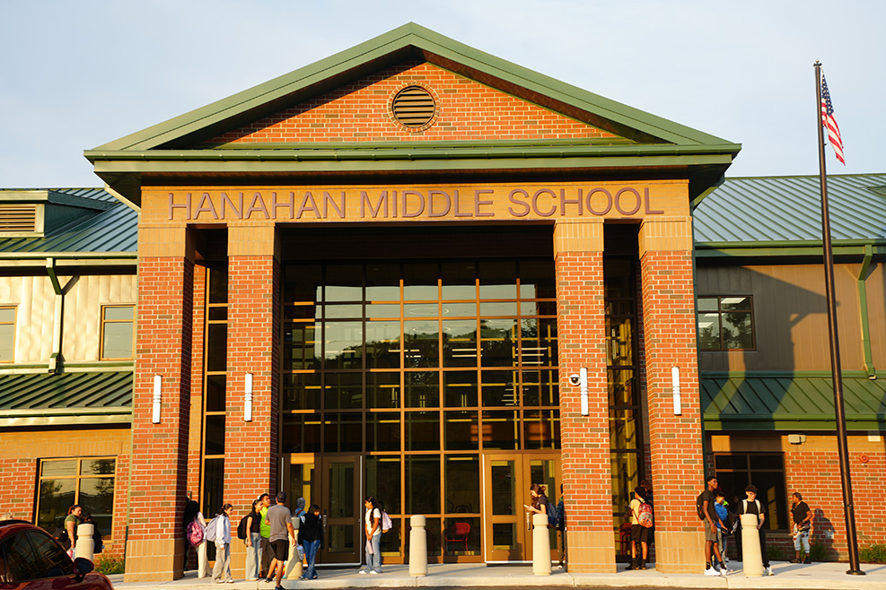 Exterior of Hanahan Middle School with brick walls, green roof, and large glass entrance. Students in front.