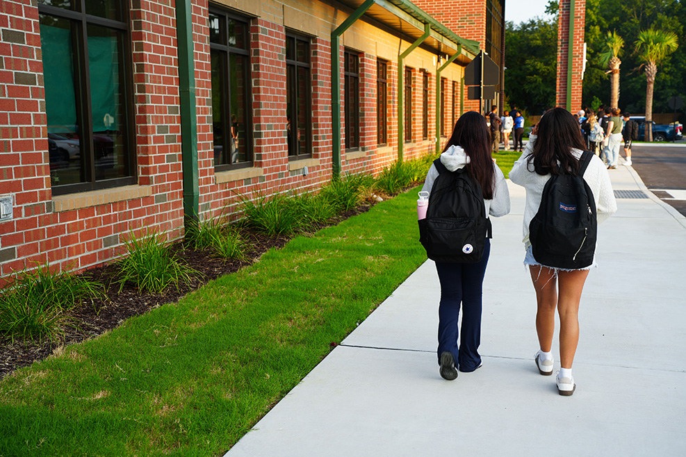 Two women walk on a sidewalk, carrying backpacks, in front of a red-brick building with large windows.
