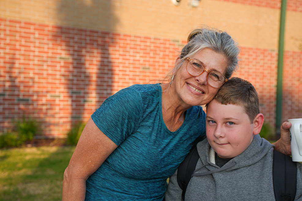 A woman and boy stand together outside with a brick building behind them. She wears glasses and a blue shirt.