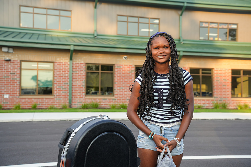 A woman in a striped shirt and denim shorts stands by a parking meter in front of a brick building.