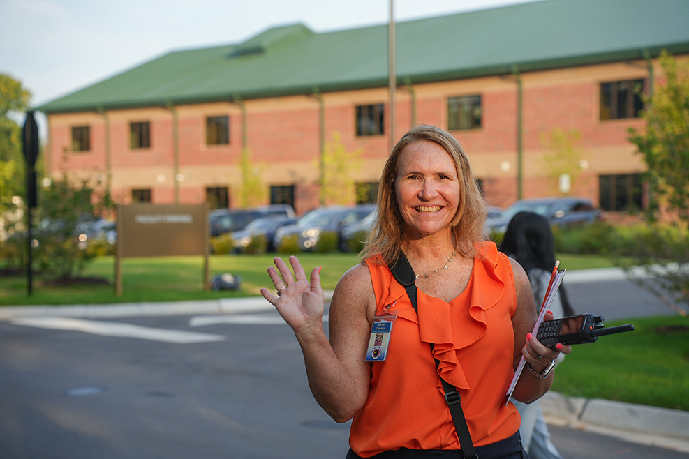 A woman in an orange top waves while holding a paper and a remote, in front of a building.