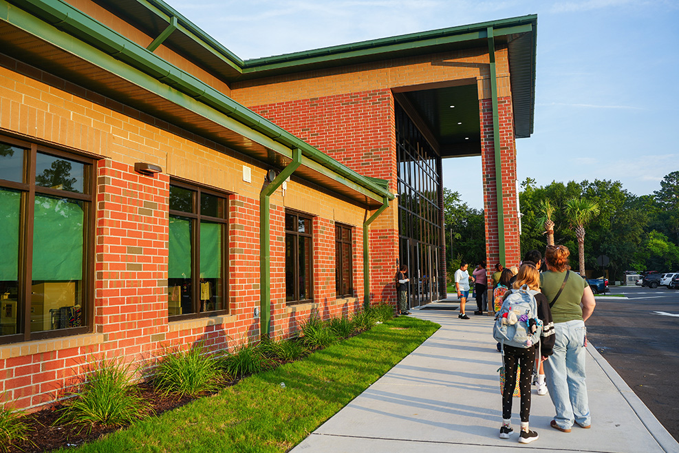 Brick building with green trim and windows. People walk on a sidewalk in front. A grassy area beside the building.