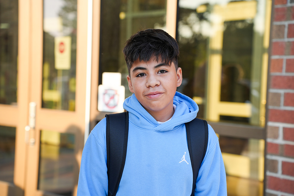 Boy with black hair, wearing a light blue hoodie, and a black backpack, standing in front of a brick wall.