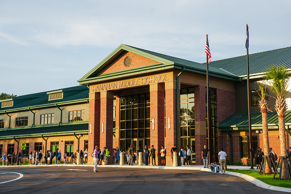 Exterior of a school building with a green roof, large windows, and several people standing outside.