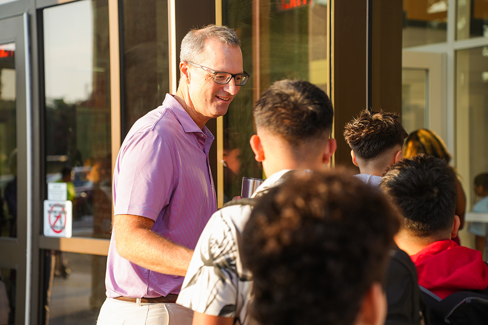 A man in glasses and a purple shirt stands near an entrance, smiling and talking to a group of people.