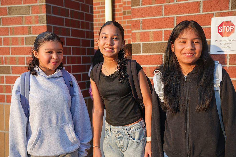 Three young women stand outside a brick wall. One wears a black shirt, another a blue hoodie, and the third a black jacket.