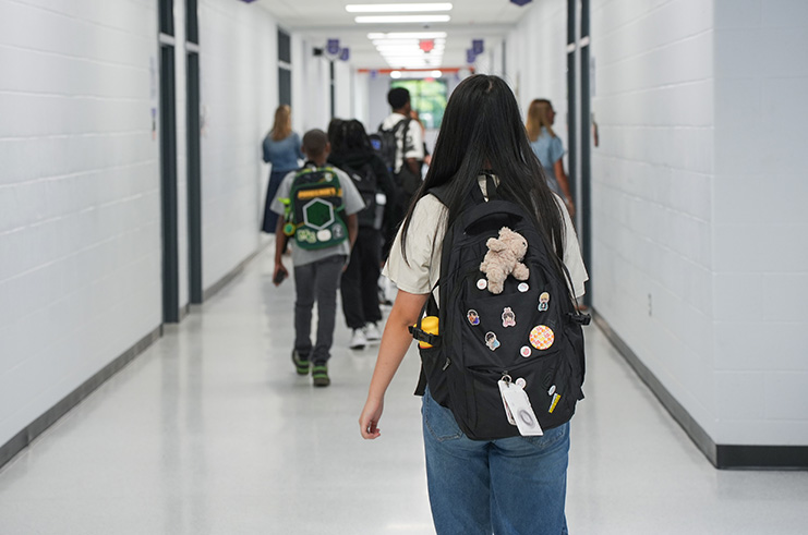 Hallway in a building with multiple people walking. One person wears a backpack with a stuffed toy.