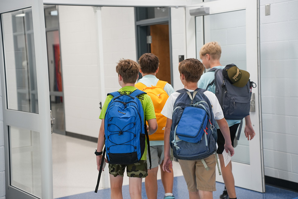 Four kids wearing backpacks and shorts walk down a hallway lined with glass doors.