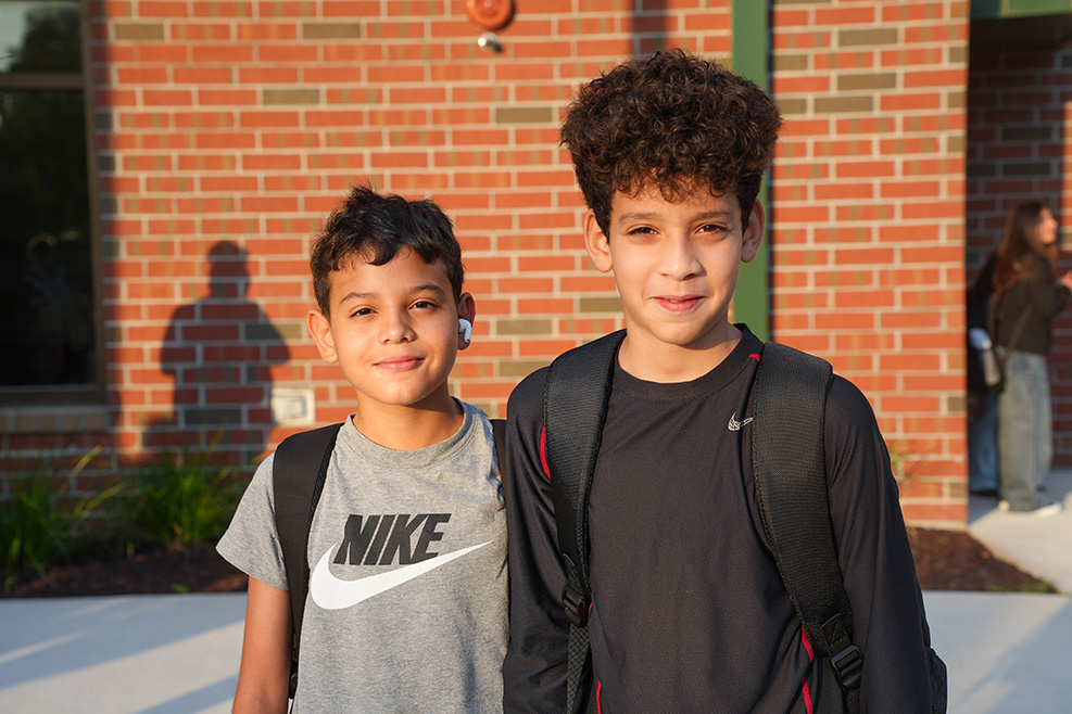 Two boys in front of a brick building. One wears a gray Nike shirt, the other a black shirt with a backpack.