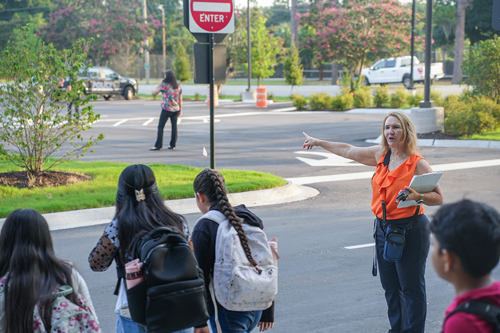 Woman in orange vest guides children with backpacks across a street; sign reads ENTER.